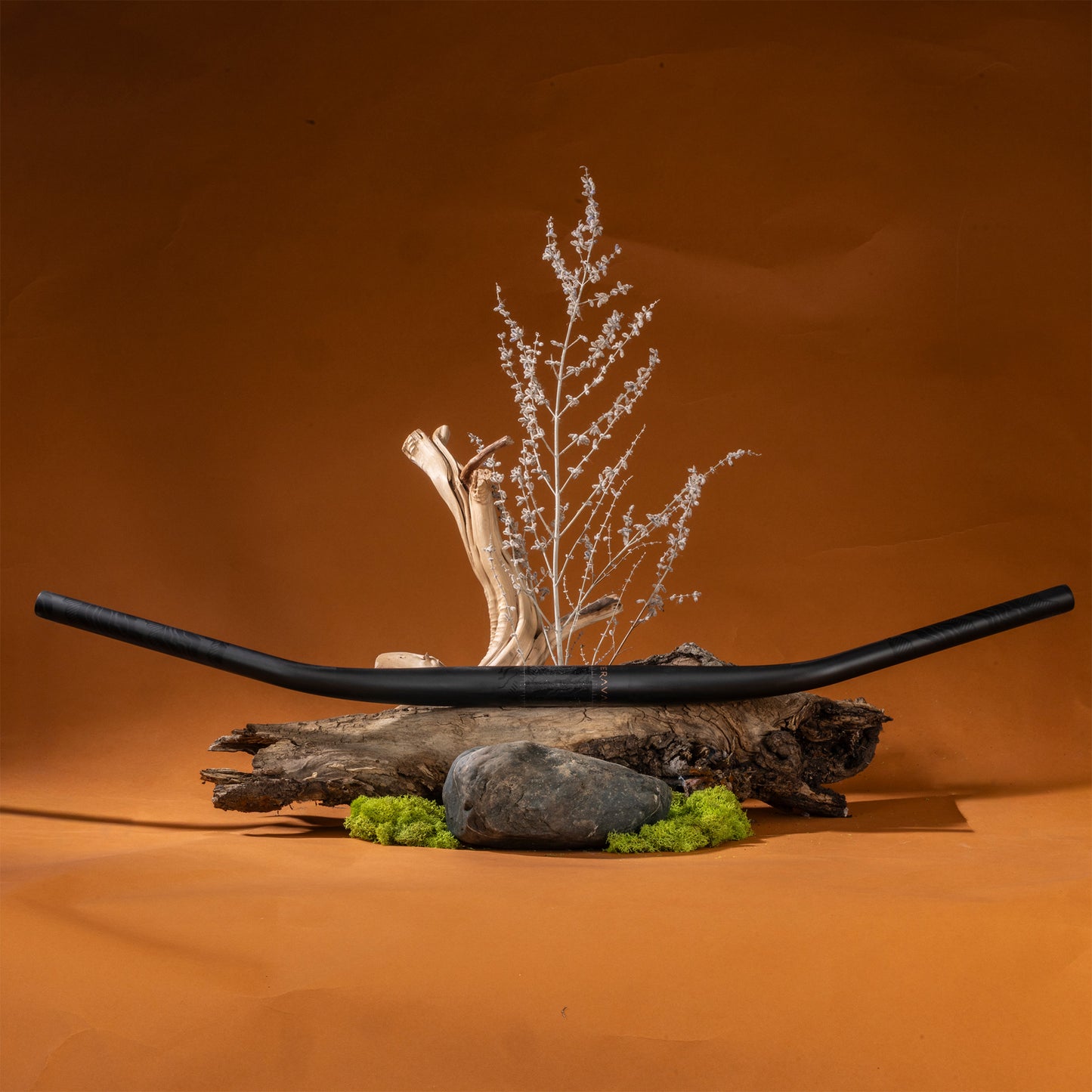 Decorative arrangement with a Swale Carbon Bar, logs, rocks, and greenery on an orange background