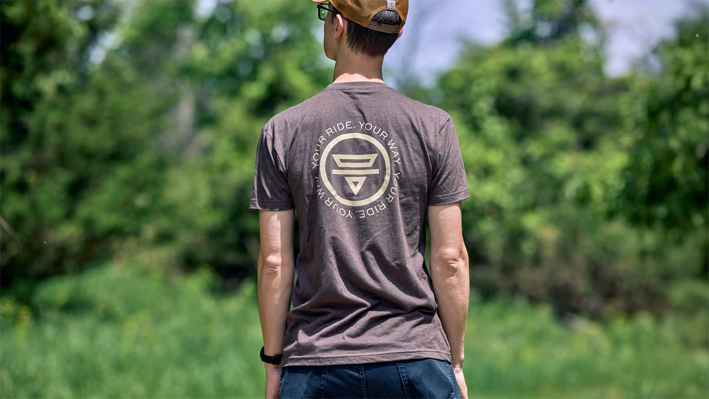Person wearing brown Teravail t-shirt with logo on back, standing outdoors with greenery in the background