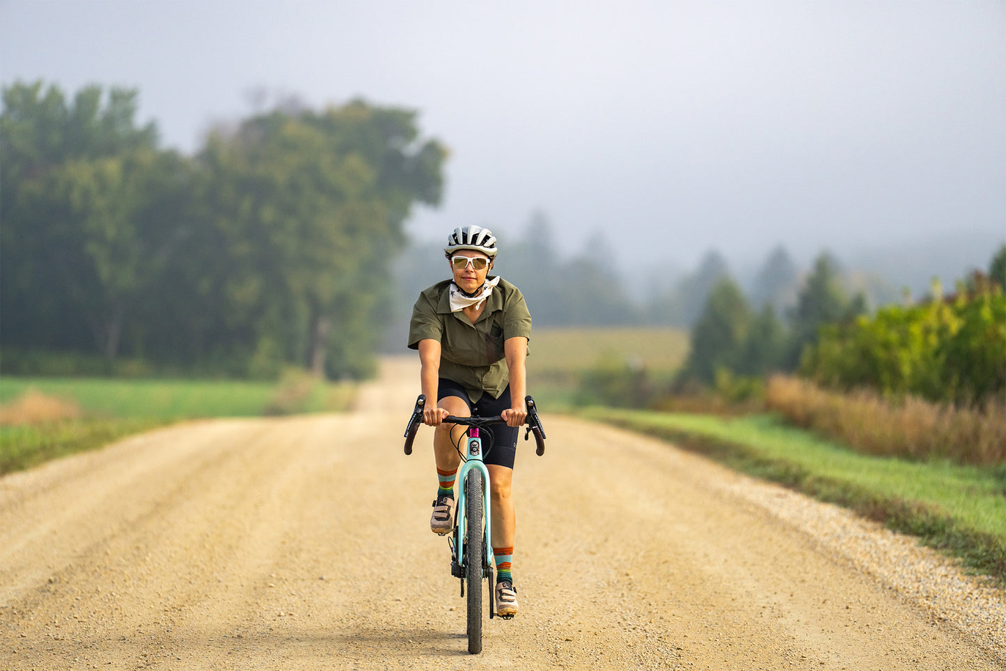 Person riding a bicycle on a dirt road with trees and fog in the background