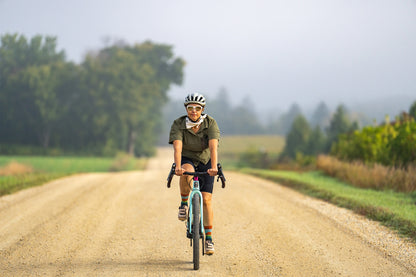 Person riding a bicycle on a dirt road with trees and fog in the background