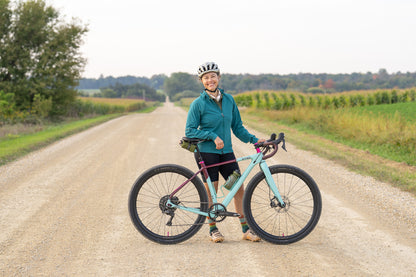 Person holding a teal bicycle on a dirt road with greenery in the background