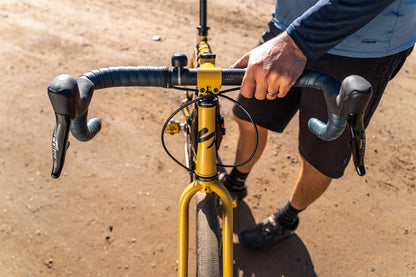 Person holding a bicycle handlebar with a yellow bike frame on a dirt road