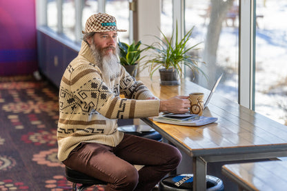 Man with a beard sitting at a table with a laptop and coffee, wearing a patterned cap and sweater.