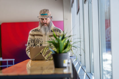 Man with a beard wearing a patterned cap and sweater, standing in an office setting with a plant and desk.