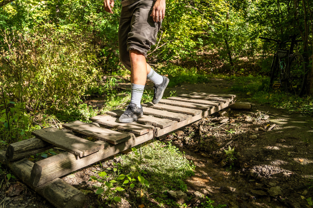 A person hiking through a forest crosses a rustic wooden plank bridge over a muddy stream, wearing dark shorts, striped socks, and hiking shoes.
