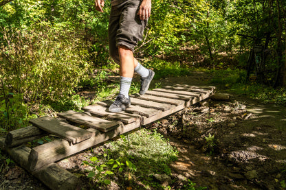 A person hiking through a forest crosses a rustic wooden plank bridge over a muddy stream, wearing dark shorts, striped socks, and hiking shoes.