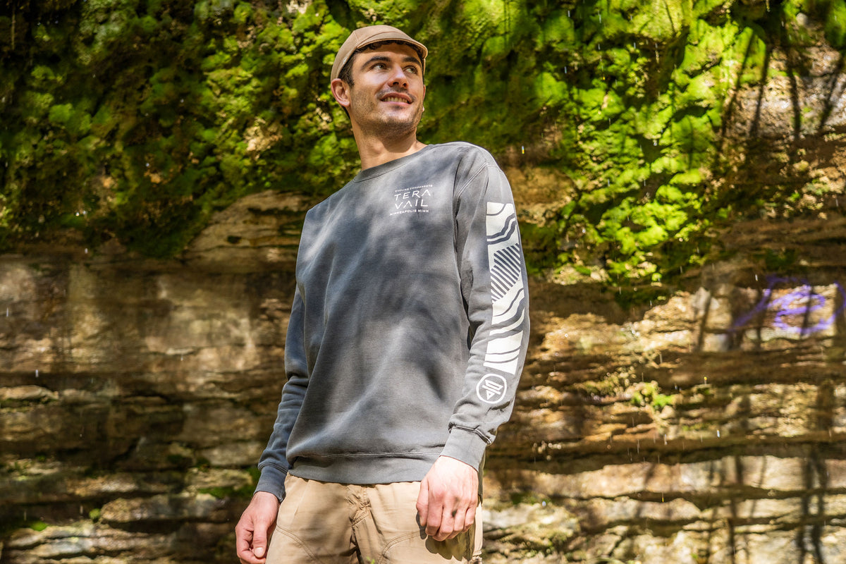 A man wearing a gray crewneck sweatshirt with a white graphic on the sleeve, khaki pants, and a tan cap, standing outdoors in front of a rocky cliff with green foliage.