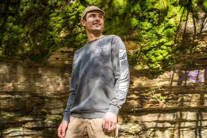 A man wearing a gray crewneck sweatshirt with a white graphic on the sleeve, khaki pants, and a tan cap, standing outdoors in front of a rocky cliff with green foliage.