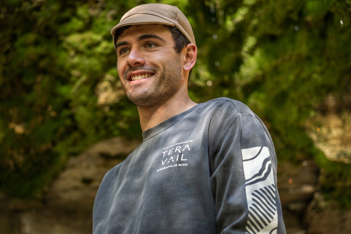 A smiling man wearing a tan cap and a dark gray long-sleeve performance shirt with 'TERAVAIL' printed on the chest, set against a blurred green forest background.
