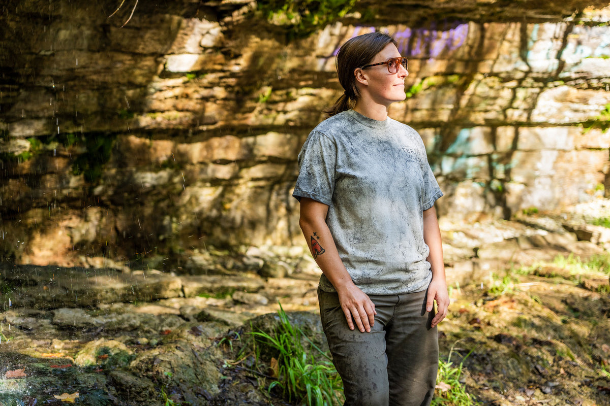 A woman wearing a light gray t-shirt and dark pants stands outdoors in front of a rocky, mossy wall, looking upward and to the side.