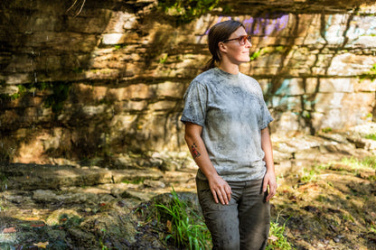 A woman wearing a light gray t-shirt and dark pants stands outdoors in front of a rocky, mossy wall, looking upward and to the side.
