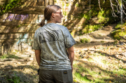 A woman wearing a Teravail short-sleeve t-shirt with a gray and black mountain graphic on the back, standing in a sunlit forest setting.