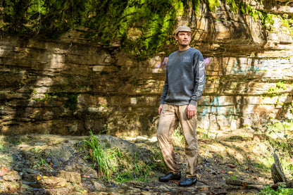 A man stands outdoors in front of a rocky cliff face with green moss and trees, wearing a gray long-sleeve crewneck shirt, tan pants, and a tan cap.