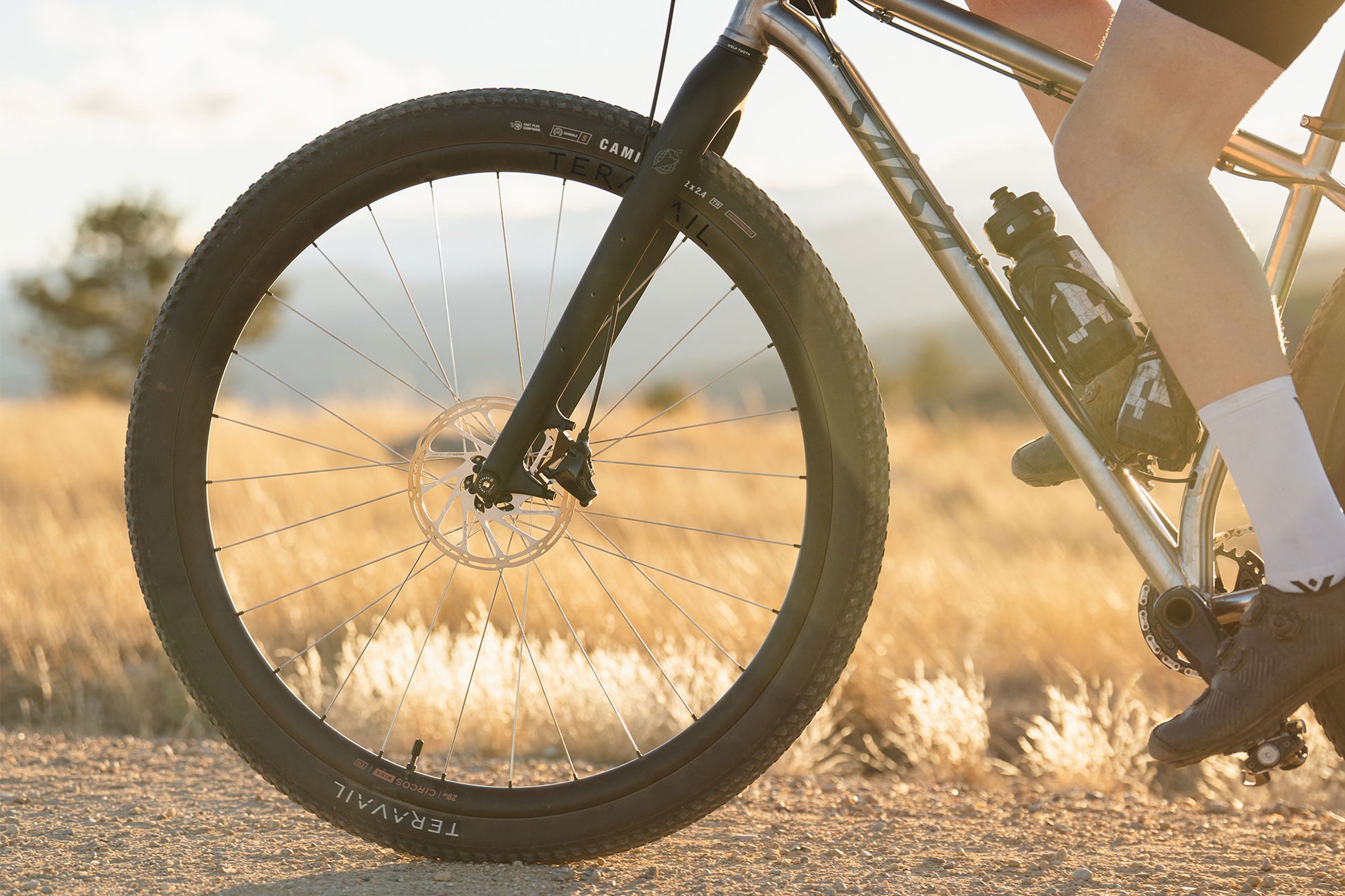 Close-up of a mountain bike wheel and front fork with a rider's legs visible, riding outdoors on a sunlit dirt path through dry grass.