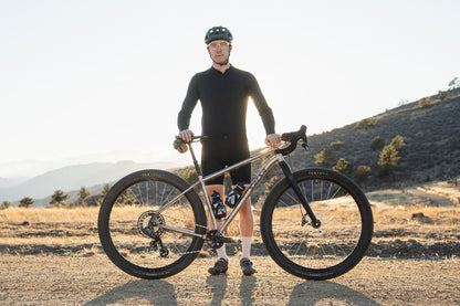A man wearing a black cycling jersey, shorts, and a helmet stands outdoors on a dirt path holding a silver gravel bicycle with black wheels, with hills in the background.