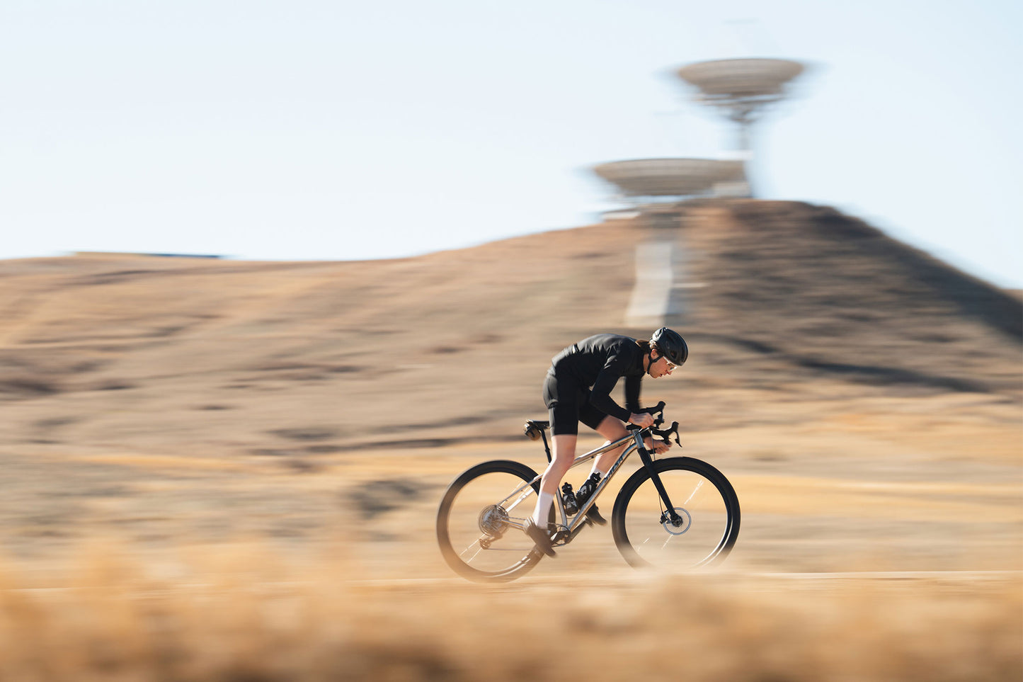 A cyclist in black cycling gear rides a road bike across a dry, grassy landscape with a blurred mountain and sky in the background.