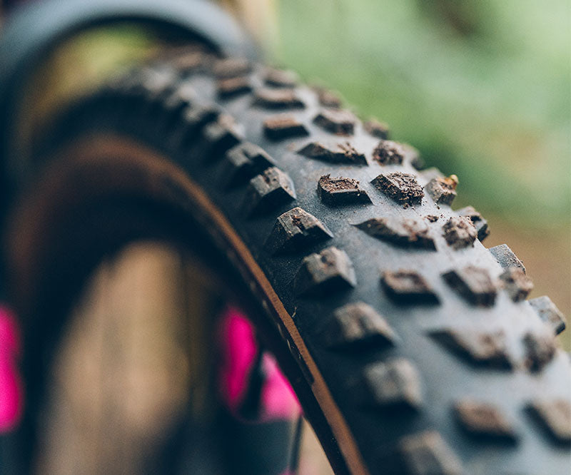 Close-up of a knobby mountain bike tire with deep tread blocks, showing dirt and mud on the rubber, photographed outdoors with a blurred green background.